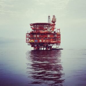 pexels photo 37703372 37703372 Dramatic view of an offshore oil rig reflecting on calm ocean waters under a cloudy sky.
